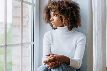 Thoughtful young woman with dark skin and modern kinky hair sits on white windowsill looking...