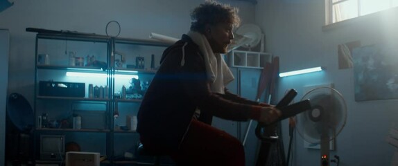 HANDHELD Portrait of unny determined overweight man in retro outfit cycling and sweating on a stationary bicycle in his home garage. Exercising during COVID-19 self isolation quarantine