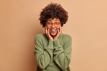 Fototapeta na wymiar Joyful woman with Afro hair keeps hands on cheeks smiles happily looks gladfully at camera wears optical glasses and sweater isolated over brown background. Positive emotions and human reactions
