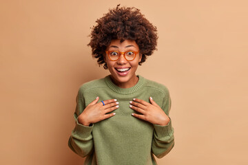 Fototapeta na wymiar Horizontal shot of happy woman keeps hands on chest and smiles gladfully reacts on getting unexpected gift wears transparent glasses and jumper isolated over brown background. Emotions concept