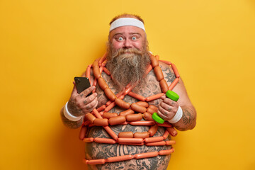 Funny plump man uses smartphone app to count how much calories he burnt sticks to diet trains in gym with dumbbell poses with sausages around naked torso. Overeating and loosing weight concept