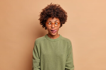 Fototapeta na wymiar Photo of Afro American teenage girl makes funny grimace crosses eyes and sticks out tongue wears basic sweater poses against brown background. Playful woman entertains child. Face expressions concept