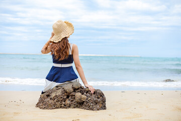 young woman barefoot ware hat and sitting on stone see wave of sea water and sand on the beach.