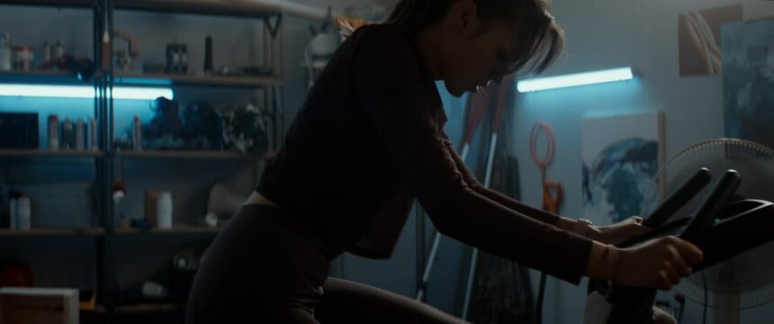 Attractive sporty Caucasian female working out on a stationary bicycle in a garage, keeping her body fit during COVID-19 coronavirus lockdown. Checking her smart fitness device
