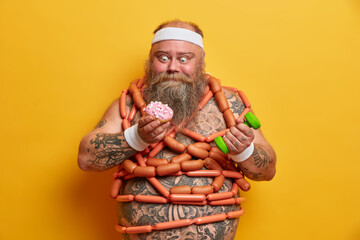 Plump bearded man obsessed with junk food tries to loose weight wears headband and wristbands has eating disorder raises dumbbell poses with naked torso wrapped body by sausages wants to eat donut