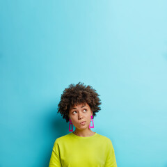 Pensive dark skinned woman purses lips and looks above thinks about something concentrated upwards...