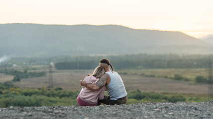 Young man hugs his girlfriend on the mountain peak at sunset background.