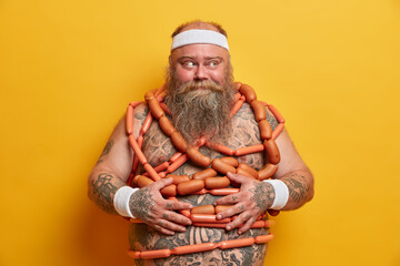 Plump bearded man with big tattooed belly wears headband and wristbands stands wrapped by sausages tries to loose weight isolated over yellow background. Stout overweight guy has bad eating habits