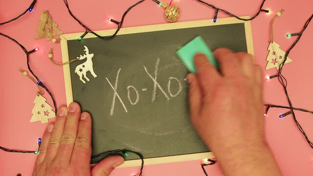 The man's hand is erased by xo xo xo on a black board and writes the end. There is an wooden toys and colored lights. Dad changes the manuscript Happy New Year greetings. Flat laid holiday items.