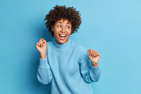 Photo of joyful dark skinned woman dances carefree keeps fists raised looks positively aside dressed in casual jumper moves against blue background. People positive emotions and feelings concept
