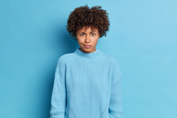 Puzzled dark skinned Afro American woman looks indignant at camera has dissatisfied expression wears knitted sweater poses against blue background hears something unexpected and bad news. Oh no
