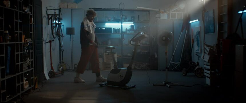 WIDE Funny overweight man in retro outfit mounting stationary bicycle in his home garage. Exercising during COVID-19 self isolation quarantine. Shot with 2x anamorphic lens 