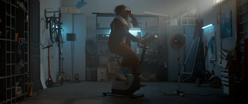 WIDE Funny overweight man in retro outfit drinking water while riding a stationary bicycle in his home garage. Exercising during COVID-19 self isolation quarantine