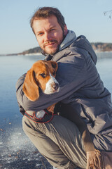 Man in winter clothes sits on shore frozen lake with a dog in his arms.
