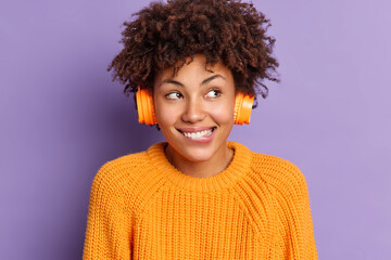 Close up shot of happy dark skinned woman listens music via headphones bites lips and enjoys...