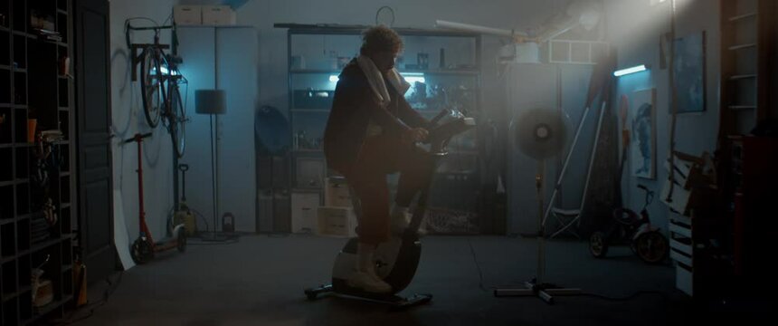 HANDHELD Funny overweight man in retro outfit dancing after riding a stationary bicycle in his home garage. Exercising during COVID-19 self isolation quarantine