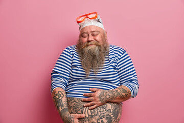 Isolated shot of plump bearded man holds his fat tattooed belly closes eyes with satisfaction dressed in undersized sailor shirt rubber swimhat and goggles enjoys snorkeling hobby during summer time