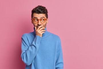 Studio shot of happy man tries to hide emotions giggles silently and covers mouth with hand looks aside positively dressed in casual clothes poses against pink background blank copy space on right
