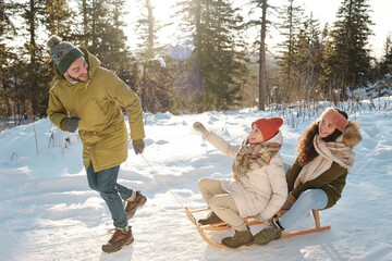 Fototapeta na wymiar Two joyful girls in winterwear sitting on sledge while young man pulling them