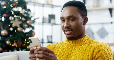 Close up portrait of joyful African American man in yellow sweater sitting at home with christmas glowing tree and texting on smartphone sending greetings and wishes to friends Happy New Year concept
