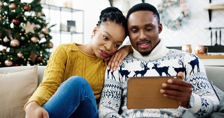 Portrait of joyful African American husband and wife sitting in modern room with decorated glowing christmas tree and tapping on tablet choosing xmas gifts online at home. Merry Christmas concept