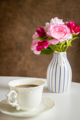 A cup of hot tea on the table behind it a stylish jug with a bouquet of flowers