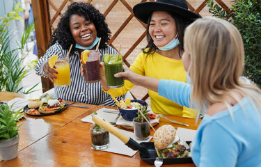 Multiracial girls have fun at brunch bar and cheering with smoothies while wearing surgical face masks under chin - Coronavirus lifestyle