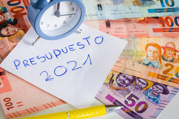 Stylish alarm clock for five to twelve. In the background a wallet with a sticking pile of Bolivian money. One hundred Boliviano banknotes