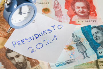 Handwritten inscription on a sheet of paper in Spanish "Presupuesto 2021" denoting the home budget in the new year. Colombian money and the alarm clock for five to twelve