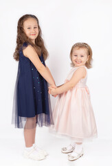 Two little beautiful smiling sister girls in beautiful dresses holding hands on a white background. isolate. A happy childhood, children's world, the concept of stylish children's clothing