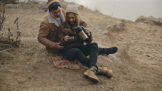Sweet young caucasian couple in love nearby autumn lake. Lovers hug and kiss having date. Woman and man have picnic, sitting on plaid and drink hot tea at cold day. Love, relationship, family concept.