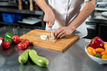 Hands of young male chef in uniform cutting fresh onions on board by table