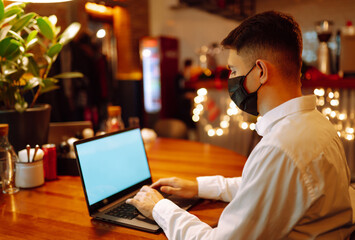 Young man with protective face mask working on laptop at home during winter holiday. Technology, bussines, freelance and remote work concept.The concept of preventing the spread of the epidemic Covid.