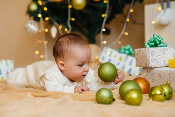 Fototapeta premium Cute smiling baby is lying under a festive Christmas tree and playing with gifts. Christmas and New Year celebrations