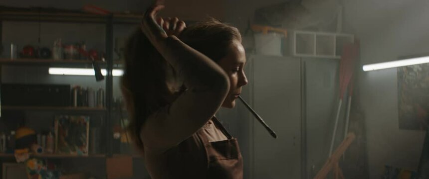 MED Female painter artist preparing to work in the studio inside her garage, ties her hair with a brush. Shot with 2x anamorphic lens