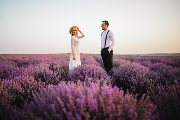 Young newlyweds dressed in country style stand in a blooming lavender field at sunset