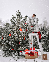 Young man in stylish silver jacket standing on stool and decorating Christmas tree with balls outdoors in winter snowy forest and beautiful nature at background. New Year mood