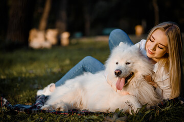 beautiful woman in white shirt is hugging her white dog samoyed outdoors in the park and sitting on the grass.