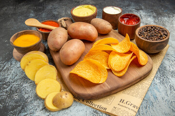 Side view of homemade delicious potato chips on wooden cutting board different spices and flavors on newspaper on gray table