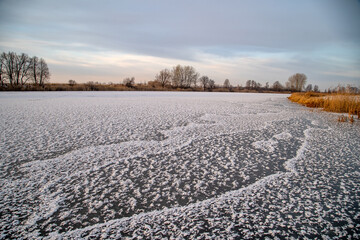 Obraz premium Frosty evening on the lake. Transparent ice with snowflakes in the form of stars. Dry, frozen grass on the shore.