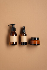 Set of amber glass dispenser bottle, sprayer and moisturizer cream jar on brown table. Flat lay, top view. Natural cosmetic products packaging design template, branding