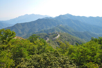Panoramic view from the majestic Great Wall of China to the surrounding green landscape and dark forests. The moutains on the horizon are in the blue haze. Trees in front in focus.