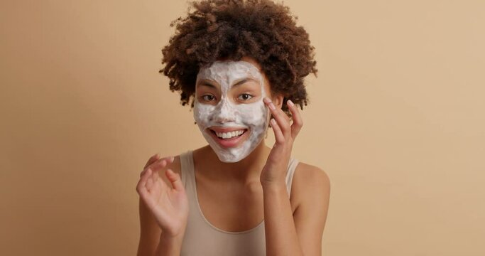 Young pretty woman with curly hair applies cosmetic product wears beauty mask on face enjoys skin care routine expresses happy emotions isolated over brown background. Facial treatment concept