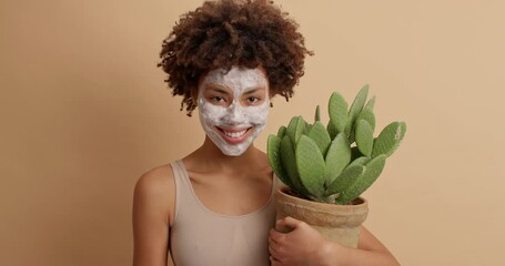 Pleasant beautiful African American woman applies nourishing face mask smoothes perfect skin has anti wrinkle procedure holds green cactus in pot poses against brown background. Grooming concept