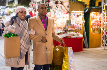 Cheerful couple with bags of gifts walking on christmas street fair