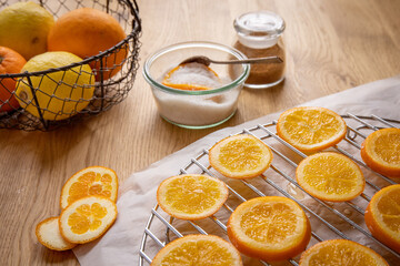 Candies Oranges Concept: A basket of citrus fruits, a bowl of sugar with an orange slice and a spoon, gingerbread spice mix, and orange slices drynig on a rack with parchment paper, on a wooden table.