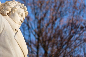 Stone figure of musical genius, composer and pianist Ludwig van Beethoven, in a monument from 1898. Horizontal daylight view with blurry leafless trees in the background and blue sky