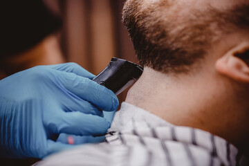Close-up of barber with medical gloves shearing beard to man in barbershop. Haircut coronavirus pandemic protection