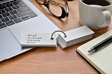 On a desk with a laptop, glasses, coffee, and a notebook, a vocabulary book was placed open There. The word Wish, Outcome, Obstacle, Plan is there.