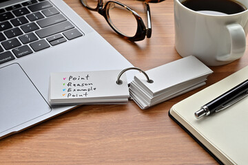 On a desk with a laptop, glasses, coffee, and a notebook, a vocabulary book was placed open There. The word Point, Reason, Example, Point is there.